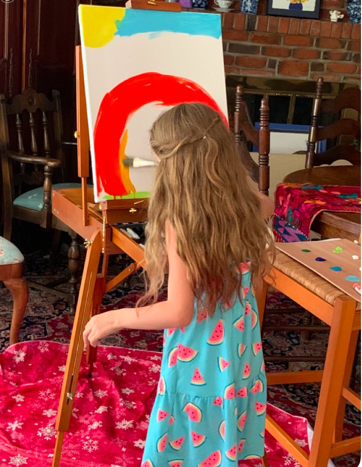 A young girl at an easel in Jean's dining room, mid-brushstroke on a bold red arc,
                paints laid out on the chair beside her